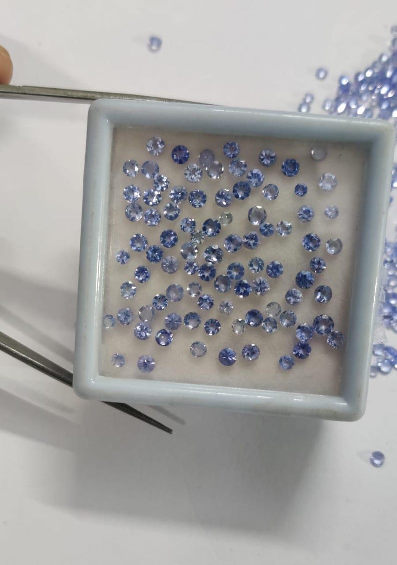 May include: A collection of small, round, light blue gemstones displayed in a white square container. The gemstones are of varying sizes and are scattered around the container. Tweezers are visible in the image, suggesting the gemstones are being handled.