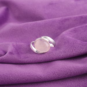 Könnte beinhalten: Ein silberner Ring mit einem großen, ovalen, hellrosa Rosenquarzstein. Der Ring ist auf einem lila Stoffhintergrund platziert. Das Band des Rings umschließt den Stein.