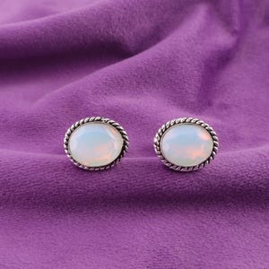May include: A pair of oval-shaped stud earrings with a silver-colored, twisted rope design around the edges. The earrings feature a milky white, opalescent stone. The background is a textured, purple fabric.