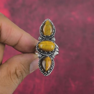Könnte beinhalten: Silberring mit drei Tigeraugensteinen in ovaler und tropfenförmiger Form. Die Steine sind gelbbraun und in eine dekorative Silberfassung mit gedrehtem Seildesign eingefasst.