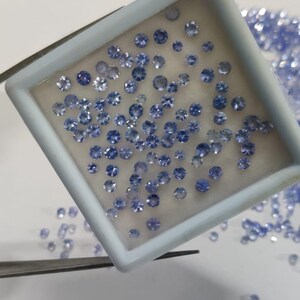 May include: A collection of small, round, light blue gemstones are displayed in a white tray. The gemstones are of varying sizes and are scattered within the tray and around it. A pair of tweezers is holding the tray.