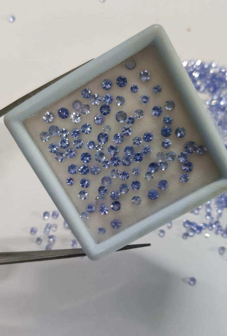 May include: A collection of small, round, light blue gemstones are displayed in a white tray. The gemstones are of varying sizes and are scattered within the tray and around it. A pair of tweezers is holding the tray.