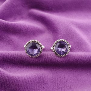 May include: A pair of silver cufflinks with a round, purple gemstone in the center. The cufflinks have a decorative, rope-like border.