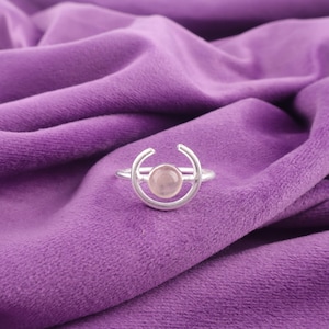 Könnte beinhalten: Ein silberner Ring mit einem Rosenquarz-Stein, der in einem Halbmond-Design gefasst ist. Der Ring wird auf einem violetten Samtstoff präsentiert. Das Design ist schlicht und elegant. Ein zarter Schmuck.