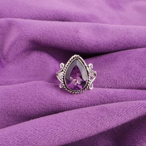 May include: A silver ring featuring a large, pear-cut purple gemstone. The gemstone is framed by a twisted rope design and two smaller, triangular white stones. The ring is set against a purple fabric background.
