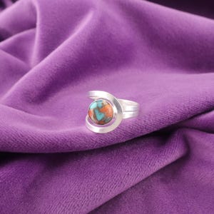 May include: A silver ring with a round, multi-colored stone featuring shades of orange, blue, and brown. The ring has a unique, open design with a curved band that wraps around the stone. The ring is displayed on a purple velvet surface.