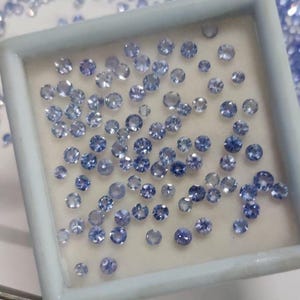 May include: A collection of small, round, light blue gemstones displayed in a square, white tray. The gemstones vary in size and are scattered across a white surface, with additional gems visible in the background.