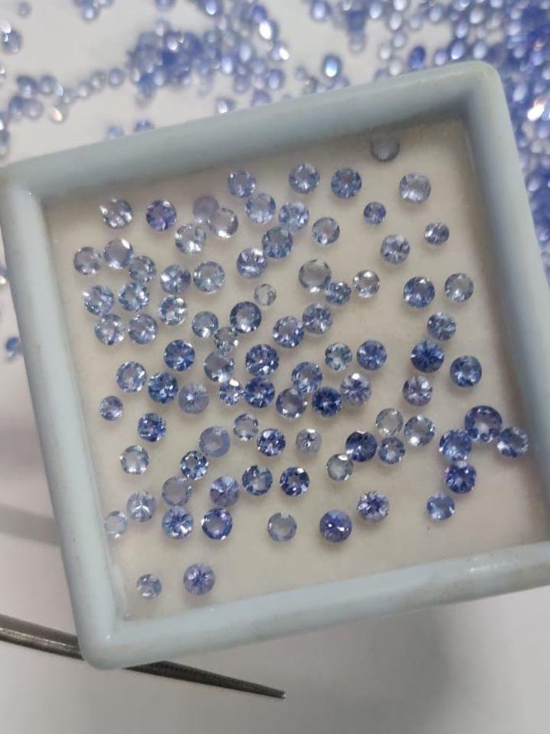 May include: A collection of small, round, light blue gemstones displayed in a square, white tray. The gemstones vary in size and are scattered across a white surface, with additional gems visible in the background.