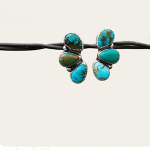 Könnte beinhalten: Ein Paar silberne Ohrringe mit Türkissteinen. Jeder Ohrring hat eine Ansammlung von drei oder vier tropfenförmigen Türkissteinen in Blau- und Grüntönen, die in einen silbernen Rahmen gefasst sind. Die Ohrringe werden vor einem weißen Hintergrund präsentiert.