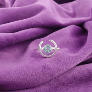 Könnte beinhalten: Ein silberner Ring mit einem hellblauen Edelstein, der in einem Halbmond-Design gefasst ist. Der Ring wird auf einem lila Samtstoff präsentiert, was einen Kontrast erzeugt.