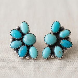 May include: A pair of silver-toned stud earrings featuring a cluster of turquoise-colored stones. Each earring has a central, round stone surrounded by smaller, oval-shaped stones, creating a floral design. The earrings are set against a neutral background.