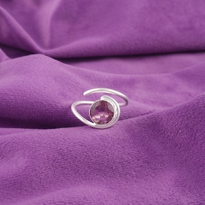 Könnte beinhalten: Ein silberner Ring mit einem großen, ovalen, purpurrosa Edelstein. Das Design des Rings zeigt ein wirbelndes Band, das sich um den Edelstein windet. Der Ring ist vor einem strukturierten, lila Hintergrund platziert.