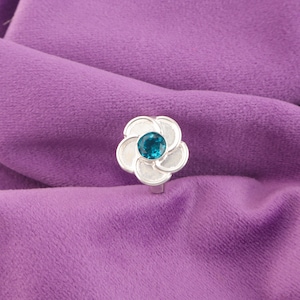 Könnte beinhalten: Ein silberner Ring mit einem Blumenmuster. Der Ring hat einen zentralen, runden, teal-farbenen Edelstein, umgeben von sechs weißen Blütenblättern. Der Ring ist vor einem lila Stoffhintergrund platziert.