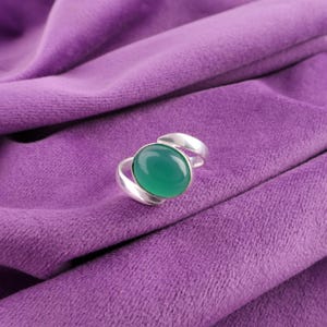 Könnte beinhalten: Ein silberner Ring mit einem großen, ovalen, leuchtend grünen Edelstein. Das Band des Rings umschließt den Stein und erzeugt ein modernes Design. Der Ring liegt auf einem tiefvioletten Stoff.