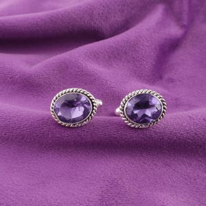 May include: Two silver rings with oval, purple gemstones. The gemstones are set in a silver frame with a twisted rope design. The rings are displayed on a purple velvet background.