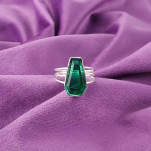 Könnte beinhalten: Ein silberner Ring mit einem großen, facettierten Edelstein in Smaragdgrün. Der Ring hat drei Bänder, die sich um den Finger wickeln.