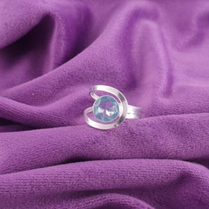May include: A silver ring with a large, light blue gemstone. The gemstone is round and faceted, set in a swirling silver band. The ring is displayed on a purple velvet surface, creating a contrast of colors and textures.