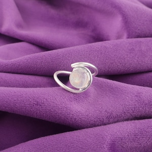 Könnte beinhalten: Ein silberner Ring mit einem runden, irisierenden Edelstein, der in einem wirbelnden Design gefasst ist. Der Ring wird auf einem violetten Samthintergrund präsentiert, der seinen eleganten und modernen Stil hervorhebt. Der Edelstein ist ein Mondstein.