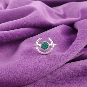 May include: A silver ring with a circular design and a vibrant green gemstone at the center. The ring's design includes a crescent shape on each side of the gemstone. The ring is displayed on a purple velvet surface.