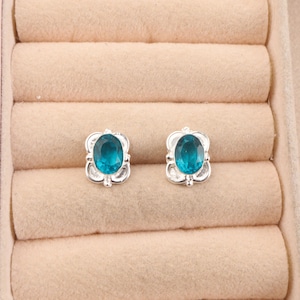 Könnte beinhalten: Ein Paar silberne Ohrringe mit blauen ovalen Edelsteinen, die in einem dekorativen Blumenmuster gefasst sind.