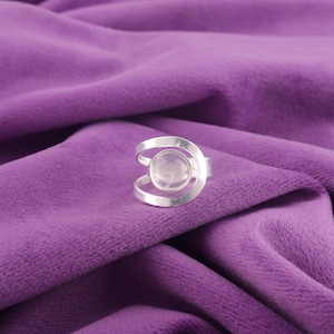 Könnte beinhalten: Ein silberner Ring mit einem klaren, runden Stein in der Mitte. Der Ring hat ein einzigartiges Design mit geschwungenen Bändern, die den Stein umschließen. Präsentiert auf einem violetten Samthintergrund.