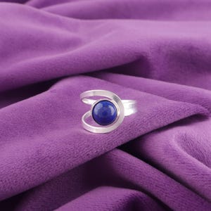 Könnte beinhalten: Ein silberner Ring mit einem wirbelnden Design, der einen runden, tiefblauen Edelstein aufweist. Der Ring ist vor einem leuchtend violetten Stoffhintergrund platziert, der den Kontrast zwischen dem kühlen Metall und der Farbe des Edelsteins hervorhebt.