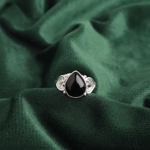 Puede incluir: Un anillo de plata con una piedra preciosa negra en forma de lágrima. El anillo tiene detalles intrincados alrededor de la piedra preciosa.