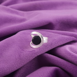 Könnte beinhalten: Ein silberner Ring mit einem großen, runden schwarzen Stein. Der Ring hat ein einzigartiges Design mit geschwungenen silbernen Akzenten, die den Stein umfassen. Vor einem violetten Samthintergrund.