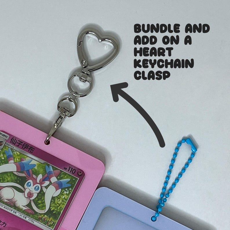 May include: A silver heart-shaped keychain clasp attached to a pink card holder featuring a Sylveon character card. The text BUNDLE AND ADD ON A HEART KEYCHAIN CLASP is printed with an arrow pointing to the clasp, next to a blue ball chain.