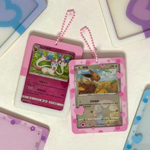 May include: Two rectangular plastic card holders with pink borders and decorative heart and flower cutouts. Each holder contains a trading card and features a pink ball chain attachment at the top. The cards display illustrated creatures and text.