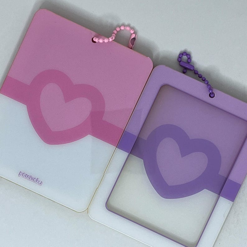 May include: Two rectangular card holders with rounded corners, one pink and one purple. Each features a central heart cutout design and a matching ball chain attached at the top. The pink holder displays the text pomixchu in the bottom corner.