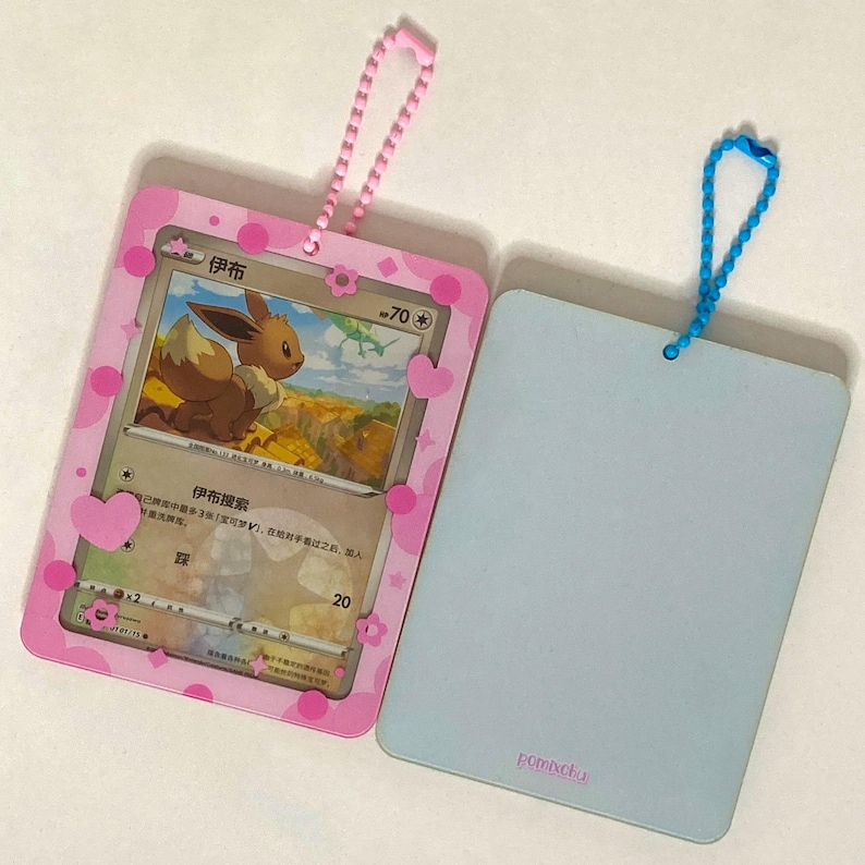May include: Two rectangular card holders with ball chain loops. One holder is pink with white and pink heart and flower accents, containing a trading card featuring a brown Eevee character. The other holder is solid light blue with a small logo.