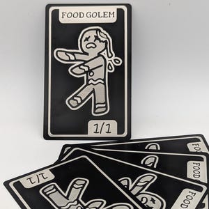Puede incluir: Cartas coleccionables en blanco y negro con un diseño estilizado de hombre de jengibre. El texto "FOOD GOLEM" y un número están presentes. Varias cartas están apiladas, mostrando el mismo diseño en diferentes orientaciones.