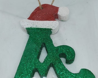 May include: Green glitter letter A Christmas ornament with a red and white Santa hat on top.