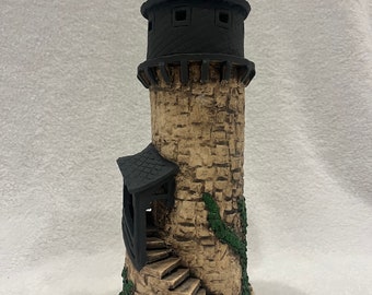 Könnte beinhalten: Ein kleiner, handgefertigter Keramikturm mit einem schwarzen Dach und einer Wendeltreppe. Der Turm ist mit einer steinartigen Textur und grünem Efeu verziert.