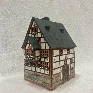 May include: A white ceramic house with a black roof and brown trim. The house has a small balcony with flowers and a door with a brown frame. The house is sitting on a white surface.