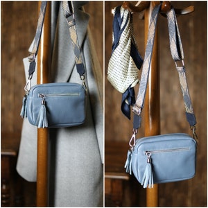 May include: A light blue leather crossbody bag with a zipper closure and two tassels. The bag has a long, adjustable strap with a geometric pattern in shades of gold and blue.