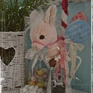 May include: A pink plush rocking horse with a white mane and tail, a blue and white striped candy cane, and a blue and white striped ball. The horse is sitting on a white pitcher with a floral design and the text "Couture" on the front. The horse is surrounded by white lace and white flowers.