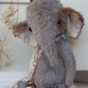 Könnte beinhalten: Ein handgefertigtes graues Elefanten-Plüschtier mit großen Ohren und grünen Augen. Der Elefant hat einen langen Rüssel und eine kleine Glocke um den Hals. Das Spielzeug ist aus weichem, strukturiertem Stoff gefertigt und hat eine Vintage-Ästhetik.