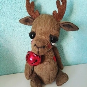 Peut inclure: Un jouet en peluche de renne brun avec de grands yeux et une cloche rouge. Le renne est assis sur une surface blanche.