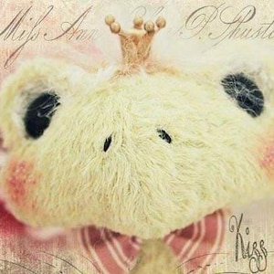 May include: A plush toy with a crown, pink cheeks, and black eyes. The toy is wearing a pink and white striped bow tie. The text "Kiss me!" is written on the background.