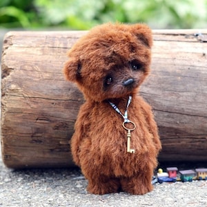 Può includere: Un orsacchiotto di peluche marrone con una chiave su un nastro blu e bianco. L'orso è in piedi su un tronco di legno.