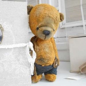 Può includere: Un orsacchiotto di peluche marrone dall'aspetto consumato, che indossa pantaloncini di jeans blu e un bottone. L'orso è in piedi su una superficie bianca.