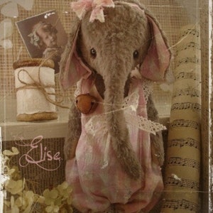 Può includere: Un giocattolo di elefante di peluche grigio che indossa un vestito rosa e bianco con un bordo di pizzo. L'elefante ha un fiocco rosa sulla testa e una campana marrone intorno al collo. Il giocattolo è seduto su uno scaffale con un rocchetto di filo e un pezzo di carta su cui è scritta la parola "Elise".