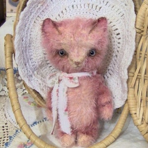 Puede incluir: Un oso de peluche rosa con un gorro de crochet blanco, que lleva una cinta floral blanca y rosa alrededor del cuello. El oso está sentado en una cesta de mimbre.