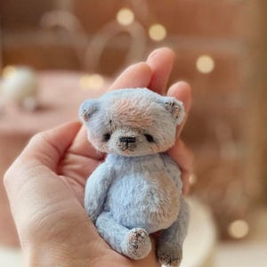 May include: A small, light blue teddy bear with a pink patch on its head. The bear is sitting in a hand.