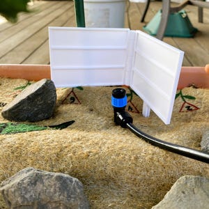May include: A white plastic fence is set up in a planter box with a brown mat and rocks. A black irrigation system with a blue connector is also present. The scene is outdoors.