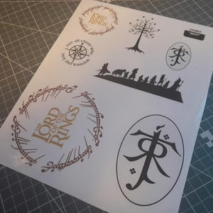 Könnte beinhalten: Ein Blatt weißes Papier mit goldenen und schwarzen Stickern zum Thema Herr der Ringe. Die Sticker enthalten die Worte "Der Herr der Ringe", einen Kompass, einen Baum und ein Symbol.