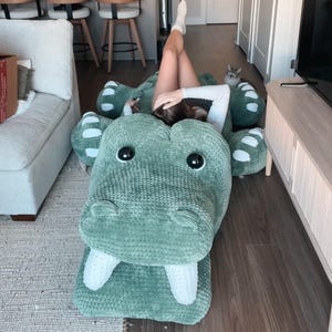 Può includere: Un grande cuscino da pavimento a forma di alligatore verde, in peluche, con denti bianchi e occhi neri. Il cuscino è realizzato in un materiale morbido e strutturato ed è progettato per rilassarsi. L'immagine è stata scattata in interni.