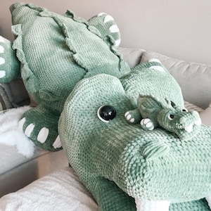 May include: A large, soft, green alligator plush toy with white accents on its feet and a smaller alligator plush. The toys are on a light gray couch with pillows and a white faux fur throw. The alligator has a textured, knitted appearance.